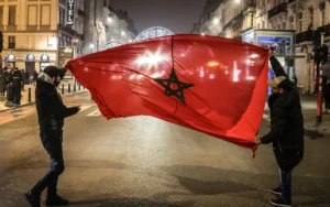 Des supporters marocains agitent joyeusement un grand drapeau rouge à l'étoile verte dans un cadre urbain animé, célébrant durant la nuit.