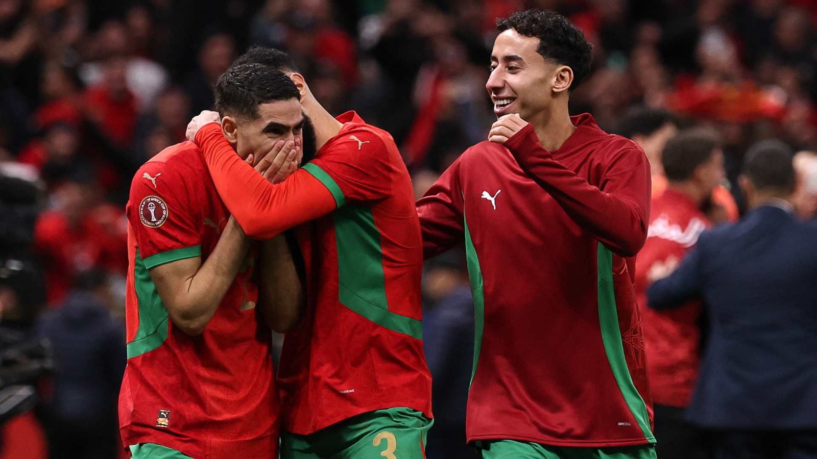 Des joueurs marocains célébrant une victoire, vêtus de tenues rouges et vertes, affichant des émotions de joie et de soulagement au sein d'une foule animée.