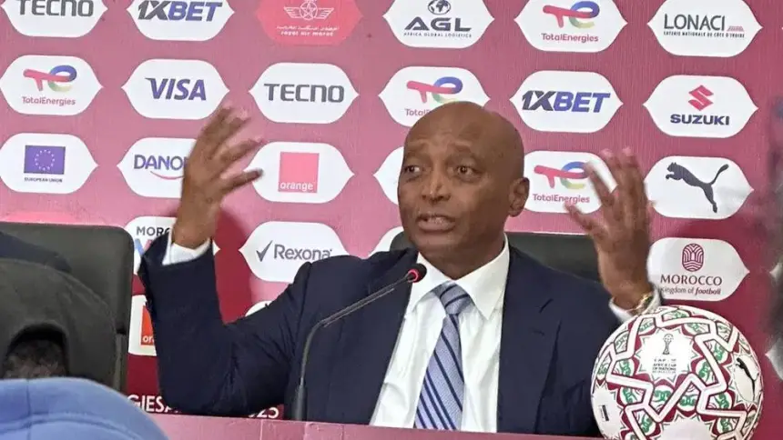 مؤتمر صحفي يظهر متحدثًا أنيقًا يشير بحماس، مع كرة قدم على الطاولة وشعارات ترويجية في الخلفية.