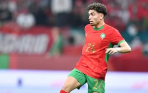 Neil El Aynaoui dans le maillot rouge et vert vif du Maroc, illustrant un mouvement dynamique lors d'un match avec des supporters enthousiastes en arrière-plan.