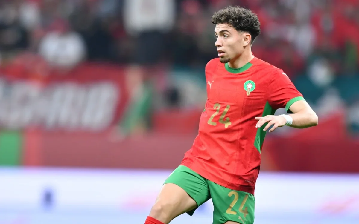 Neil El Aynaoui dans le maillot rouge et vert vif du Maroc, illustrant un mouvement dynamique lors d'un match avec des supporters enthousiastes en arrière-plan.