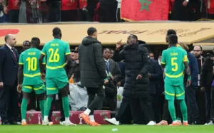 Senegalese spelers Mané en Guéye converseren met teamgenoten en staf tijdens een wedstrijd, gekleed in groen-gele tenues, met een Marokkaanse vlag zichtbaar op de achtergrond.
