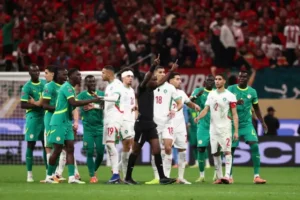 Morocco and Senegal players in heated discussion with the referee during a tense moment in a match, set against a vibrant crowd backdrop.