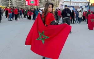 Un supporter marocain brandit fièrement le drapeau national orné d'une étoile verte au sein d'une foule animée, en célébration à l'extérieur du stade pendant un match.