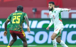 Noussair Mazraoui en tenue blanche et verte du Maroc défend contre Dina Ebimbe du Cameroun, qui porte un maillot vert vif avec des accents rouges.