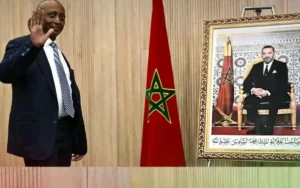 Patrice Motsepe salue à côté d'un portrait du roi Mohammed VI, avec un drapeau marocain en arrière-plan, symbolisant le leadership de la CAF au Maroc.