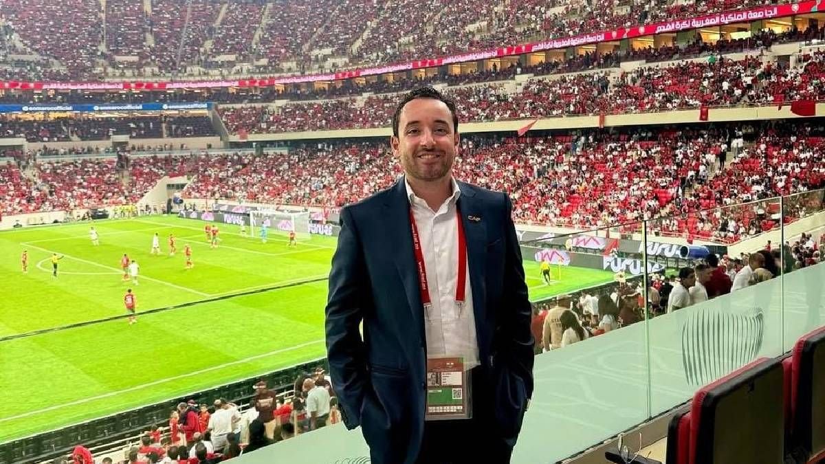 A man in a suit stands in a stadium, smiling, with hundreds of fans in red seats behind him and players on the pitch during a match.