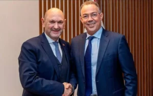 Rafael Louzan en een Marokkaanse voetbalfunctionaris schudden elkaar de hand in een formele setting, beiden gekleed in pak, terwijl ze naar de camera glimlachen.