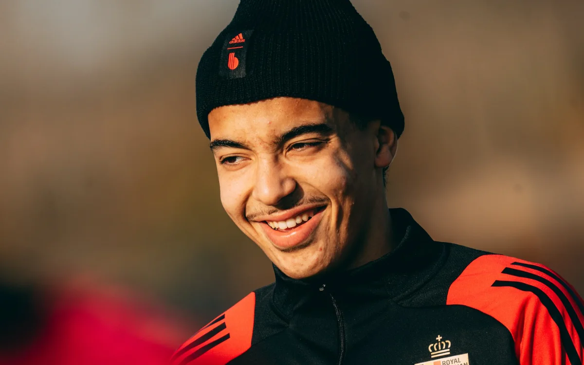 Rayane Bounida smiling during a training session, wearing a black beanie and red-accented sportswear, in a warm lighting setting.