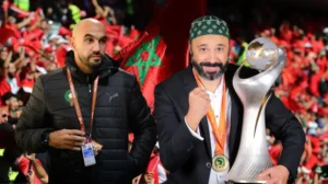 Walid Regragui se tient fièrement avec le trophée de la CAF, célébrant le succès du Maroc, tandis qu'un stade rempli de supporters agite des drapeaux marocains en arrière-plan.