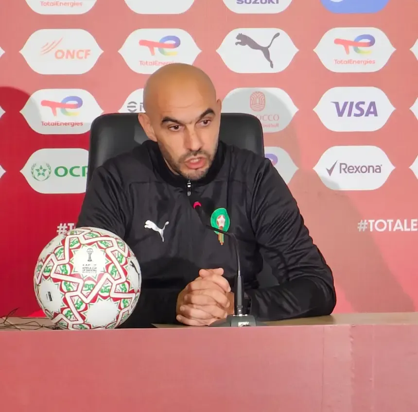 Walid Regragui speaking at a press conference, wearing a black jacket with a Morocco badge, a football at the table, with sponsor logos in the background.