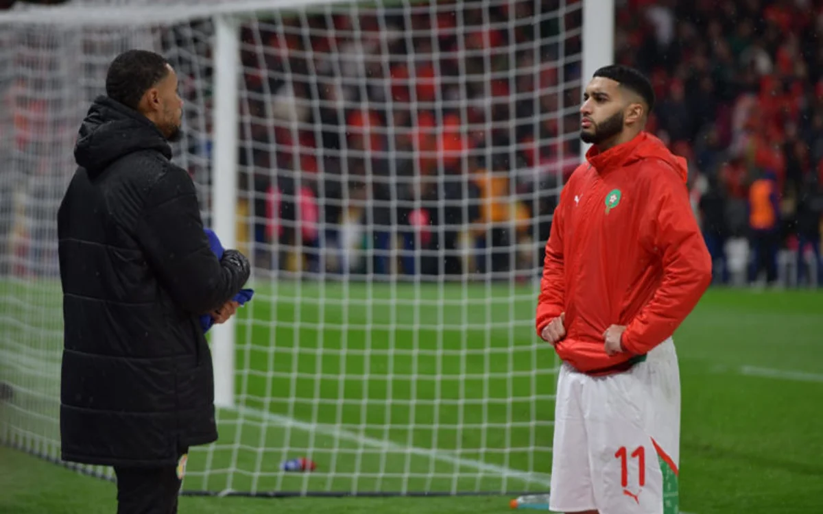 Saibari and Hakimi in Moroccan kit, deep in conversation on the pitch, with the goalpost and crowd blurred in the background.