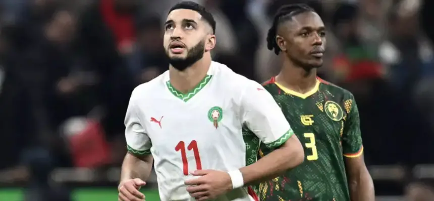 Morocco's player wearing jersey number 11, with a white and green kit, looks up thoughtfully as a Cameroonian player in green stands nearby.