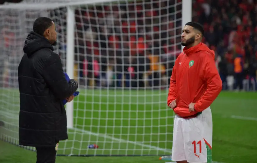 Saïbari, vêtu de la veste rouge et du short de l'équipe nationale marocaine, se tient pensivement près du but, s'entretenant avec un entraîneur.