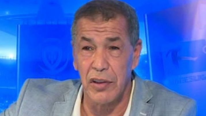 Analyste de football masculin avec des cheveux gris courts portant un blazer bleu clair, parlant sur un fond bleu à thème sportif.