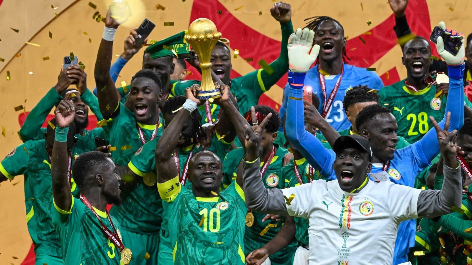 Les joueurs sénégalais célèbrent avec le trophée africain, vêtus de tenues vertes et de médailles, au milieu des confettis et avec des expressions joyeuses.