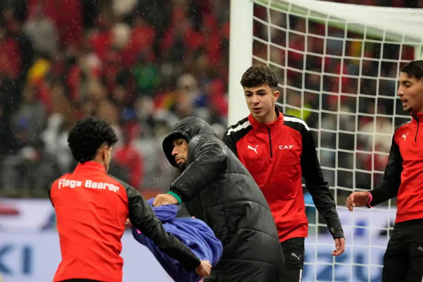 Moroccan team officials assist players in red CAF jackets on a rainy day, preparing for an upcoming match amid a lively stadium backdrop.