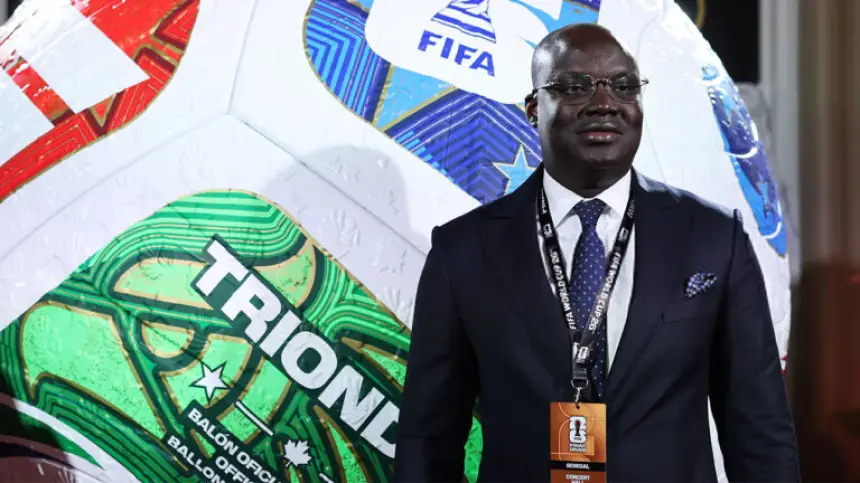 Man in a suit with glasses stands beside a large FIFA-branded football, featuring vibrant green and blue patterns, at a promotional event.