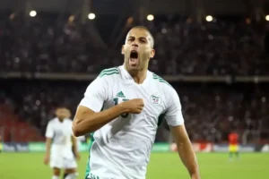 Algerian player celebrating passionately in a white kit with green accents, showcasing pride after a goal, with a crowded stadium background.