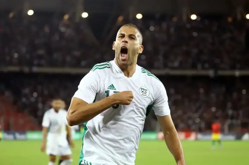Algerian player celebrating passionately in a white kit with green accents, showcasing pride after a goal, with a crowded stadium background.