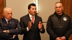 Trois hommes en tenue professionnelle discutent ; l'un fait des gestes tandis que les autres écoutent attentivement, reflétant une conversation sérieuse liée au football.