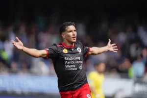 Sohayb viert vreugdevol in een zwart-rood tenue na het scoren, met een levendige stadionachtergrond en opgewonden fans.