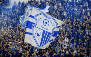Ittihad Tangier supporters wave a large blue and white flag featuring the club's logo, creating a vibrant atmosphere in the stadium.