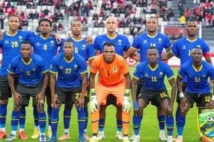 Tanzania national football team posing for a team photo in blue and yellow kits, with the goalkeeper in orange, against a stadium backdrop.