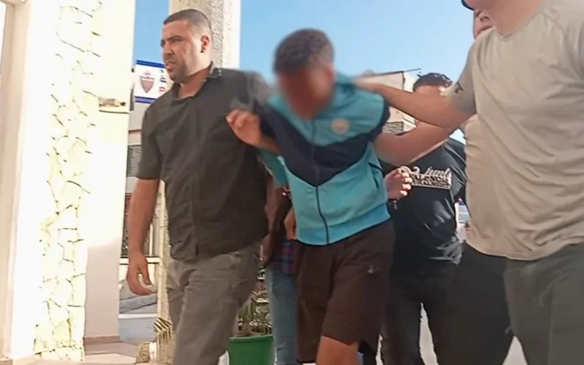 Un jeune garçon en survêtement bleu est escorté par deux hommes dans une rue ensoleillée, avec des bâtiments et des personnes en arrière-plan.