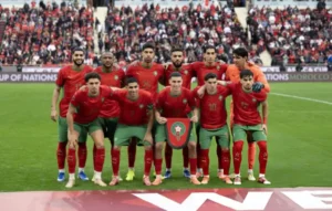 Morocco's national football team poses for a photo before a match, wearing red and green kits, with fans in the background.