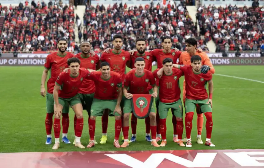 Morocco's national football team poses for a photo before a match, wearing red and green kits, with fans in the background.