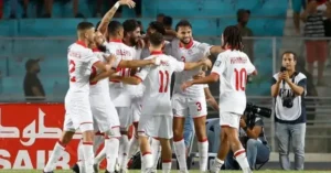 لاعبو المنتخب التونسي يحتفلون بتسجيل هدف، مرتدين زيهم الأبيض والأحمر، مع خلفية الملعب وجماهير تهتف في المدرجات.