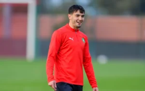 Un joueur marocain sourit pendant l'entraînement dans un survêtement rouge vif, montrant l'esprit d'équipe avant un match contre le Cameroun.