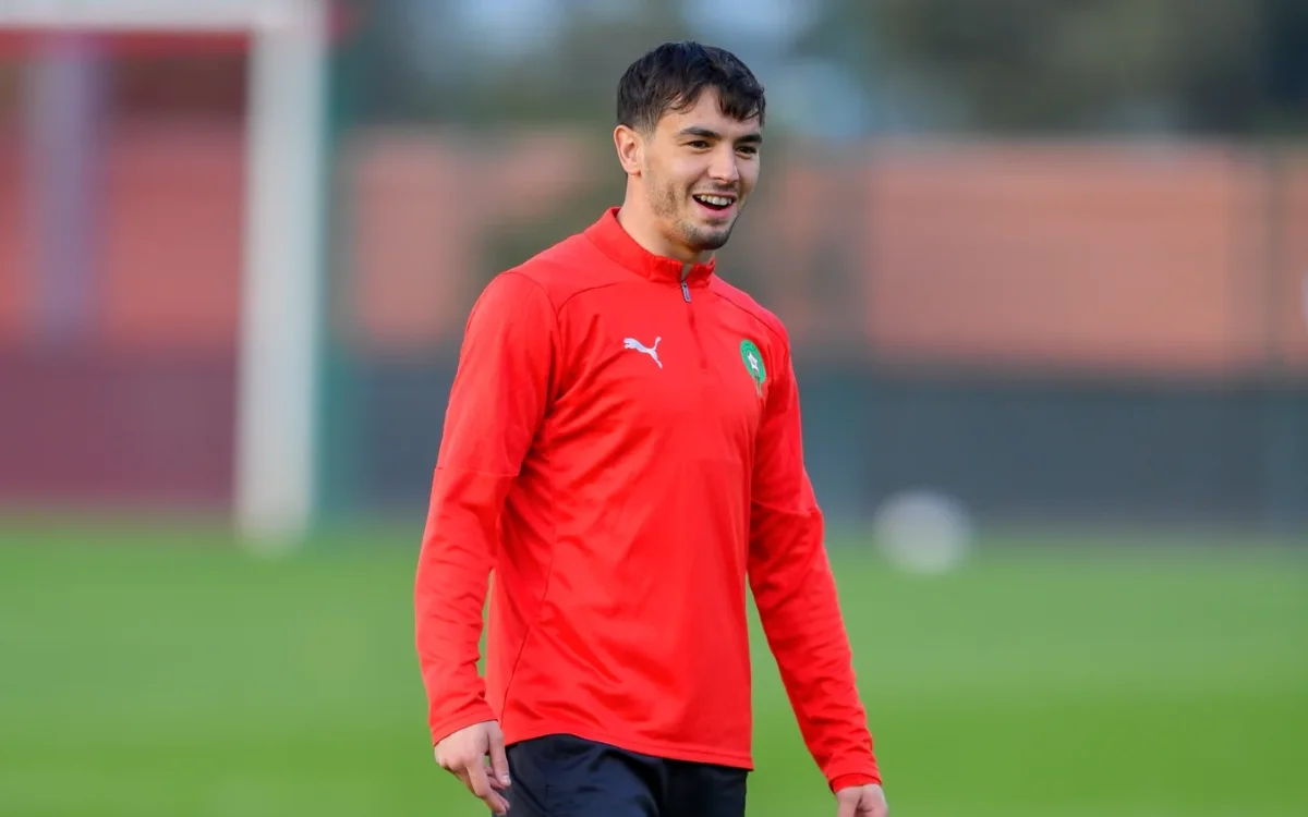 Un joueur marocain sourit pendant l'entraînement dans un survêtement rouge vif, montrant l'esprit d'équipe avant un match contre le Cameroun.