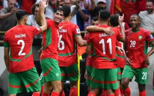 Moroccan players celebrate a goal, wearing their vibrant red and green kits, with notable players Hakimi and Saïss in focus, during a match.
