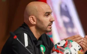 Walid Regragui speaks passionately during a press conference, with a Moroccan flag and a football adorned with national colours in view.