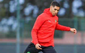 Achraf Hakimi training in a red Morocco tracksuit, focused on the ball, with a blurred background of a training facility.