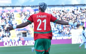 Yassir Zabiri célèbre les bras grands ouverts, portant le maillot vert et rouge de l'équipe nationale marocaine, numéro 21, dans une atmosphère vibrante du stade.