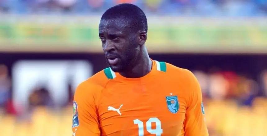 Yaya Touré en maillot orange de la Côte d'Ivoire, l'air pensif sur le terrain pendant un match, avec des supporters et le stade en arrière-plan.