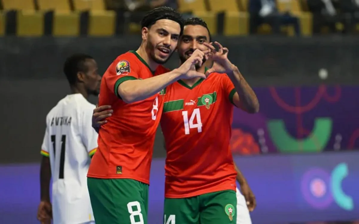 Les joueurs marocains de futsal célèbrent un but en formant un geste de cœur, vêtus de tenues rouges et vertes, avec un adversaire flou en arrière-plan.