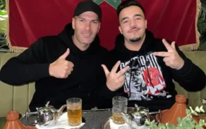 Zinedine Zidane and a fan pose happily with thumbs up and peace signs, sitting at a Moroccan-style table with tea and a tagine, against a Moroccan flag backdrop.