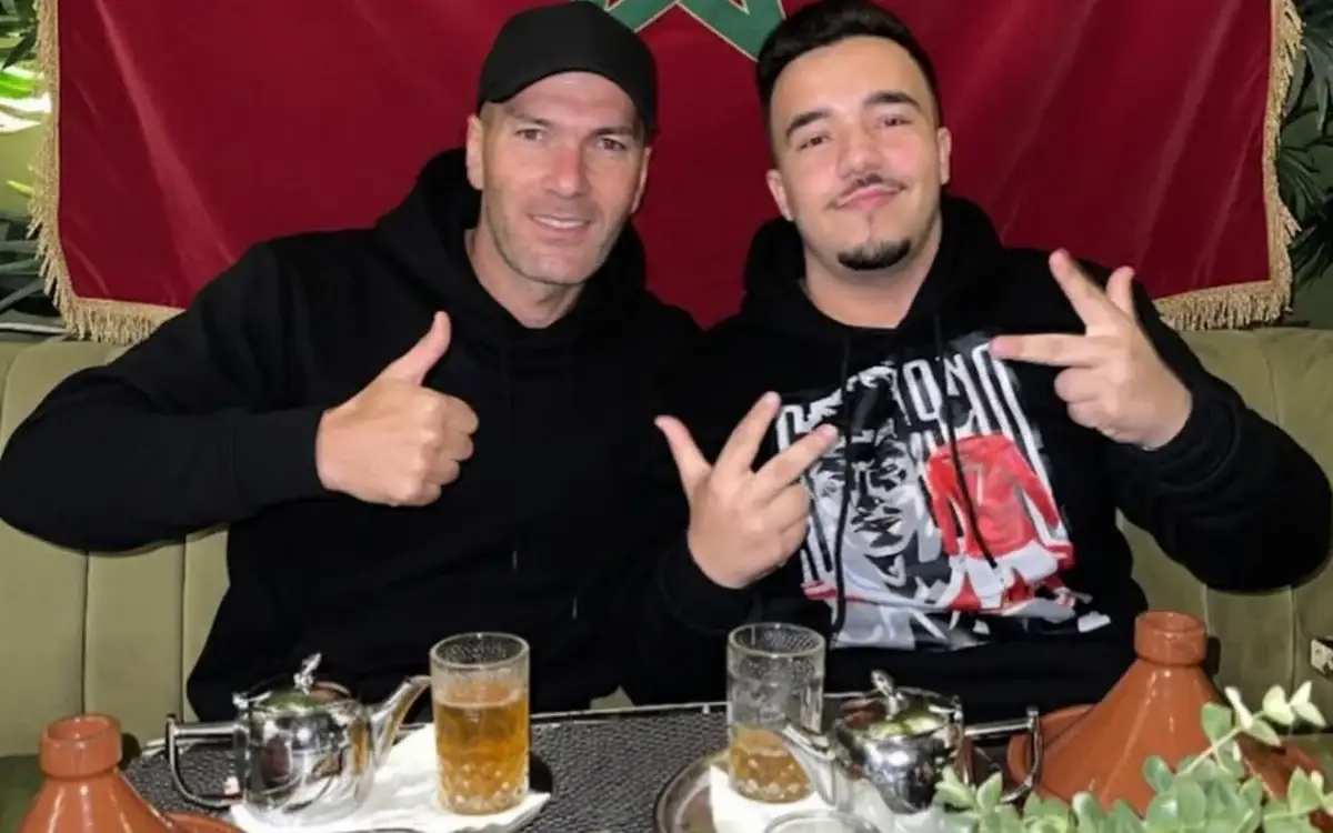 Zinedine Zidane and a fan pose happily with thumbs up and peace signs, sitting at a Moroccan-style table with tea and a tagine, against a Moroccan flag backdrop.