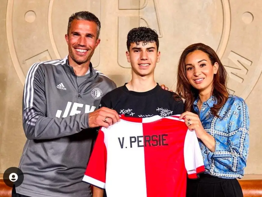 Robin van Persie lacht naast een jonge speler die een rood-wit tenue vasthoudt met "V. PERSIE," terwijl een vrouw in een blauw gestreept shirt glimlacht.