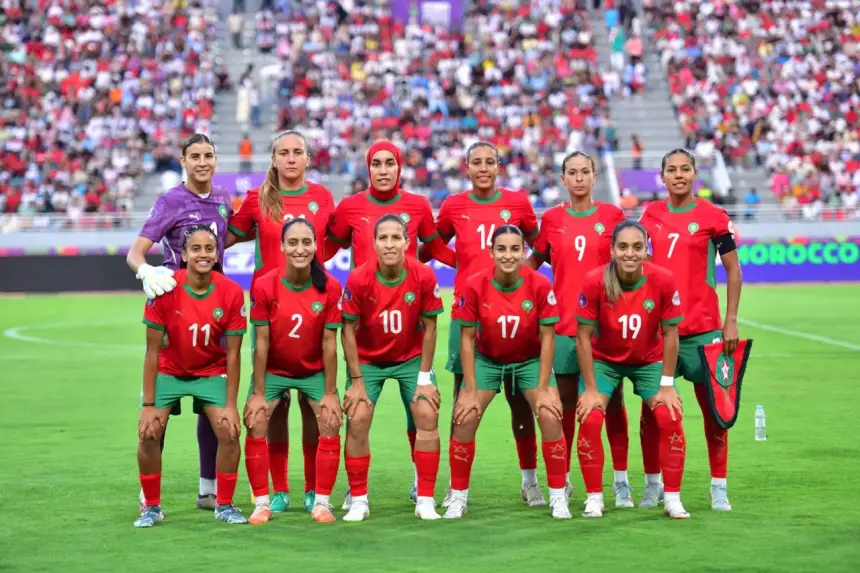 Morocco's women's national football team poses together in vibrant red and green kits, with a stadium crowd in the background.