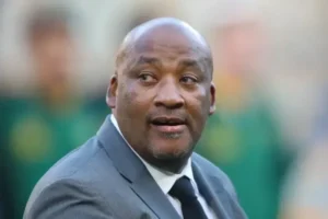 Entraîneur de football en costume gris, légèrement tourné, absorbé par l'ambiance du match. L'arrière-plan vert flou évoque le cadre d'un stade.