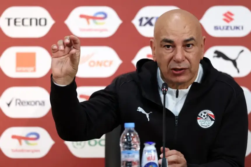 Hossam Hassan gesticule lors d'une conférence de presse devant des décors publicitaires, en discutant des stratégies footballistiques égyptiennes.