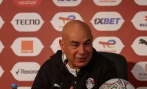 Hossam Hassan lors d'une conférence de presse, veste noire arborant le logo de l'équipe nationale égyptienne, souriant devant un fond de toile de fond publicitaire.