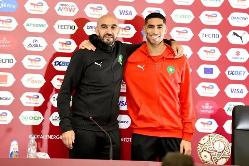 Achraf Hakimi and coach Walid Regragui pose together at a press conference, showcasing Moroccan team spirit against a vibrant backdrop.