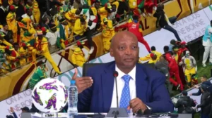 Ahmed Ahmed, voormalig CAF-voorzitter, spreekt op een persconferentie, terwijl fans op de achtergrond feestvieren tijdens een Afrikaans toernooi.