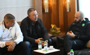 Trois hommes en tenue décontractée engagés dans une conversation, assis dans un cadre moderne avec des boissons sur une table, illustrant l'esprit d'équipe dans le football marocain.